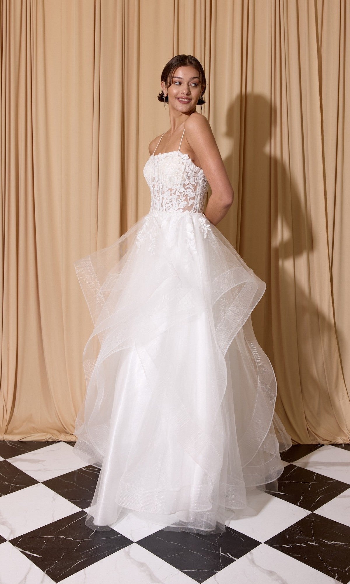 A woman in the Eureka EK167 long prom dress—white, strapless, with a glitter tulle A-line skirt and floral lace embroidered corset—stands on a black-and-white checkered floor before beige curtains.